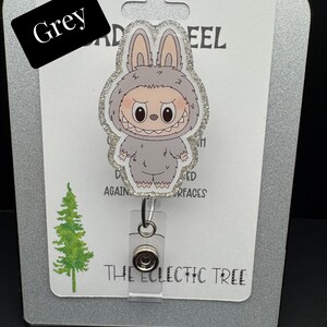 Puede incluir: Un carrete de insignia gris con un monstruo de dibujos animados con orejas de conejo. El carrete de la insignia tiene un borde brillante y un carrete retr&aacute;ctil de pl&aacute;stico transparente. La palabra "Grey" est&aacute; en un rect&aacute;ngulo negro. El texto "THE ECLECTIC TREE" est&aacute; en la parte inferior.