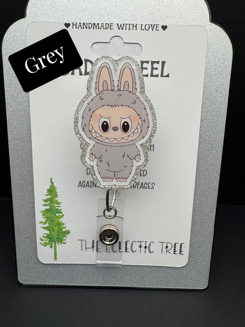 Puede incluir: Un carrete de insignia gris con un monstruo de dibujos animados con orejas de conejo. El carrete de la insignia tiene un borde brillante y un carrete retr&aacute;ctil de pl&aacute;stico transparente. La palabra "Grey" est&aacute; en un rect&aacute;ngulo negro. El texto "THE ECLECTIC TREE" est&aacute; en la parte inferior.