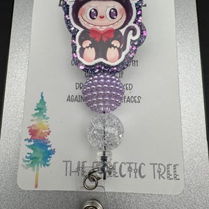 May include: A retractable badge reel featuring a cartoon cat character in a black cat costume with a red bow tie. The reel has purple and clear decorative beads and the text "THE ECLECTIC TREE".