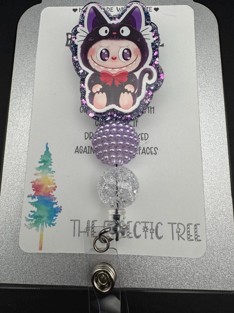 May include: A retractable badge reel featuring a cartoon cat character in a black cat costume with a red bow tie. The reel has purple and clear decorative beads and the text "THE ECLECTIC TREE".