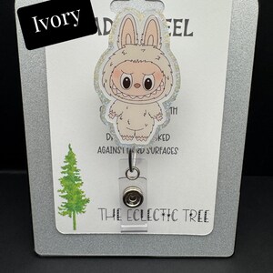 Puede incluir: Porta-credenciales de color marfil con un personaje de yeti de dibujos animados con una expresi&oacute;n de mal humor. El porta-credenciales tiene un clip de pl&aacute;stico transparente y un cord&oacute;n retr&aacute;ctil. El texto "HANDMADE WITH LOVE" y "THE ECLECTIC TREE" son visibles.
