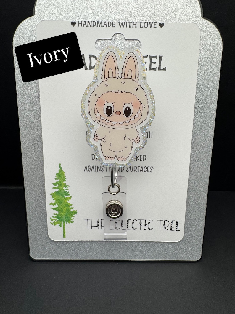 Puede incluir: Porta-credenciales de color marfil con un personaje de yeti de dibujos animados con una expresi&oacute;n de mal humor. El porta-credenciales tiene un clip de pl&aacute;stico transparente y un cord&oacute;n retr&aacute;ctil. El texto "HANDMADE WITH LOVE" y "THE ECLECTIC TREE" son visibles.