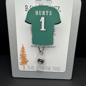 May include: A green jersey-shaped badge reel with the name "HURTS" and the number "1" printed on it. The badge reel is attached to a clear plastic holder with a retractable cord. The background includes text that reads "HANDMADE WITH LOVE" and "THE ECLECTIC TREE."