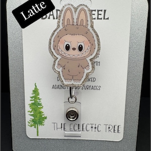 Puede incluir: Un portatarjetas con un personaje de dibujos animados con un disfraz de conejito y un borde brillante. El portatarjetas tiene un soporte de pl&aacute;stico transparente y un clip met&aacute;lico. El texto "HANDMADE WITH LOVE" y "THE ECLECTIC TREE" son visibles.