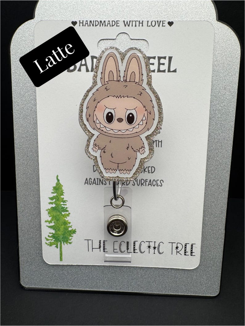 Puede incluir: Un portatarjetas con un personaje de dibujos animados con un disfraz de conejito y un borde brillante. El portatarjetas tiene un soporte de pl&aacute;stico transparente y un clip met&aacute;lico. El texto "HANDMADE WITH LOVE" y "THE ECLECTIC TREE" son visibles.