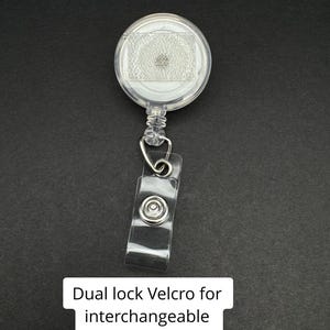 May include: A clear, round retractable badge reel with a clear plastic strap and metal clip. The reel has a dual lock Velcro for interchangeable toppers. The text "Dual lock Velcro for interchangeable toppers" is displayed on a white rectangle.