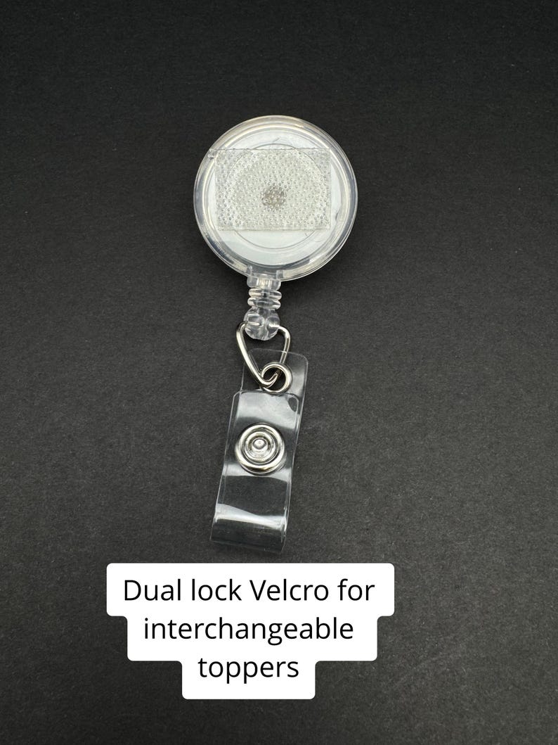 May include: A clear, round retractable badge reel with a clear plastic strap and metal clip. The reel has a dual lock Velcro for interchangeable toppers. The text "Dual lock Velcro for interchangeable toppers" is displayed on a white rectangle.