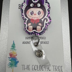May include: A badge reel featuring a cartoon character in a black cat costume with a red bow tie. The badge reel has a glittery purple border and a clear plastic strap. The text "THE ECLECTIC TREE" is printed at the bottom.