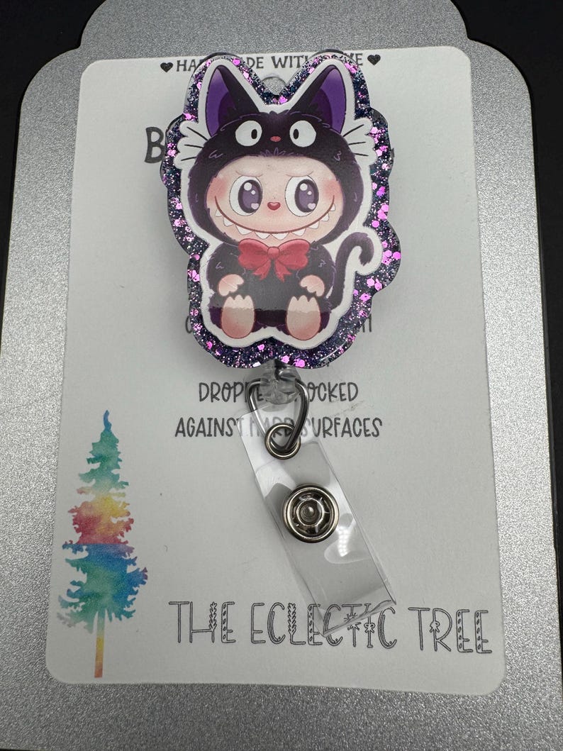 May include: A badge reel featuring a cartoon character in a black cat costume with a red bow tie. The badge reel has a glittery purple border and a clear plastic strap. The text "THE ECLECTIC TREE" is printed at the bottom.