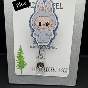 Puede incluir: Un portatarjetas con un personaje de dibujos animados con un disfraz de conejito azul con un borde brillante. El portatarjetas tiene un soporte de pl&aacute;stico transparente y un cord&oacute;n retr&aacute;ctil. Las palabras "HANDMADE WITH LOVE" est&aacute;n en la parte superior. El texto "Blue" est&aacute; en una caja negra.