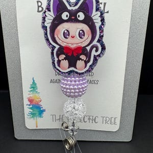 May include: A badge reel featuring a cartoon character in a black cat costume with a red bow tie. The badge reel has a purple and clear beaded design and the text "HANDMADE WITH LOVE" at the top.