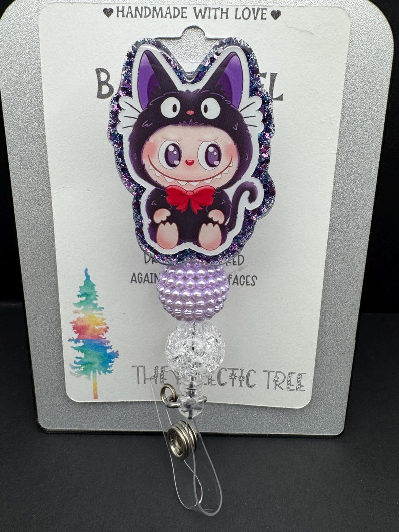 May include: A badge reel featuring a cartoon character in a black cat costume with a red bow tie. The badge reel has a purple and clear beaded design and the text "HANDMADE WITH LOVE" at the top.