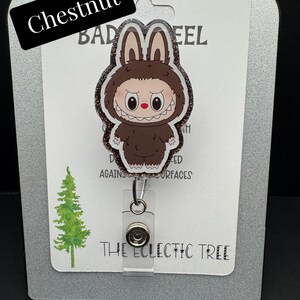 Puede incluir: Un carrete de insignia con una criatura marr&oacute;n de dibujos animados con orejas de conejo y una sonrisa amenazante. El carrete de la insignia est&aacute; etiquetado "Chestnut" e incluye el texto "THE ECLECTIC TREE". El fondo incluye un gr&aacute;fico de &aacute;rbol verde.