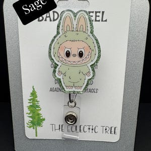 Puede incluir: Un portatarjetas verde salvia con un monstruo de dibujos animados con un disfraz de conejito y detalles brillantes. El portatarjetas tiene un cord&oacute;n retr&aacute;ctil de pl&aacute;stico transparente y un clip met&aacute;lico. El texto "Sage" est&aacute; en una etiqueta negra. El texto "THE ECLECTIC TREE" est&aacute; en la parte inferior.