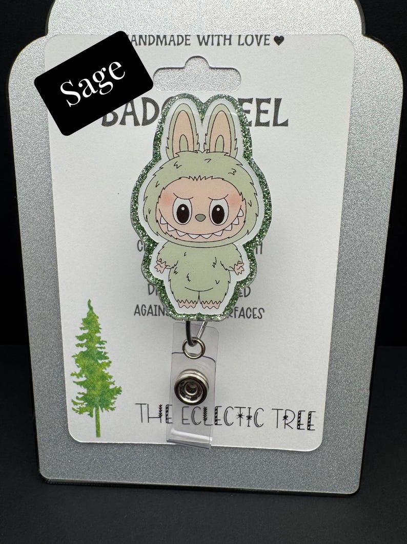 Puede incluir: Un portatarjetas verde salvia con un monstruo de dibujos animados con un disfraz de conejito y detalles brillantes. El portatarjetas tiene un cord&oacute;n retr&aacute;ctil de pl&aacute;stico transparente y un clip met&aacute;lico. El texto "Sage" est&aacute; en una etiqueta negra. El texto "THE ECLECTIC TREE" est&aacute; en la parte inferior.