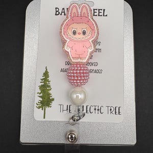 Puede incluir: Un carrete de insignia rosa con un personaje de dibujos animados con disfraz de conejito. El carrete tiene un acento de cuentas y una perla blanca. El texto "THE LECTIC TREE" est&aacute; impreso en la tarjeta.