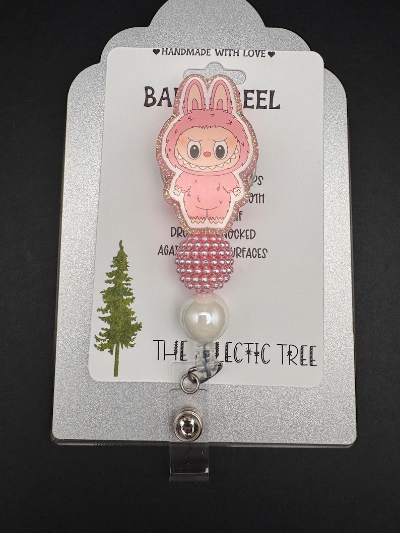 Puede incluir: Un carrete de insignia rosa con un personaje de dibujos animados con disfraz de conejito. El carrete tiene un acento de cuentas y una perla blanca. El texto "THE LECTIC TREE" est&aacute; impreso en la tarjeta.