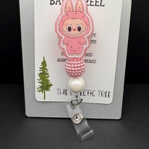 Puede incluir: Un carrete de insignia rosa con un personaje de dibujos animados con disfraz de conejo. El carrete tiene un acabado brillante y est&aacute; unido a un acento de cuentas blancas y rosas. El texto "HANDMADE WITH LOVE" y "THE ECLECTIC TREE" son visibles.