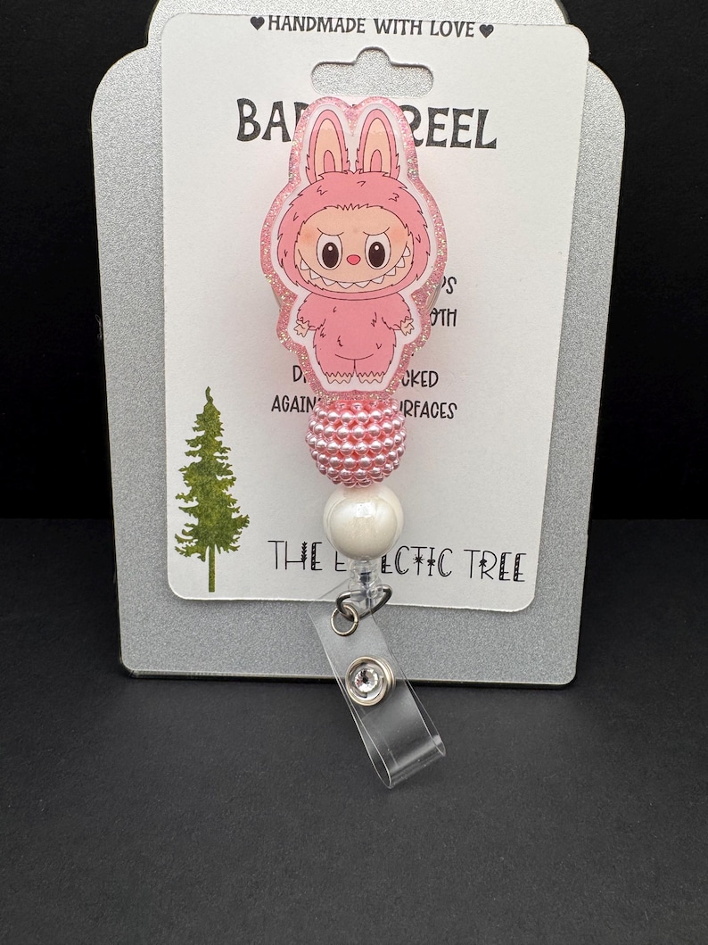 Puede incluir: Un carrete de insignia rosa con un personaje de dibujos animados con disfraz de conejo. El carrete tiene un acabado brillante y est&aacute; unido a un acento de cuentas blancas y rosas. El texto "HANDMADE WITH LOVE" y "THE ECLECTIC TREE" son visibles.