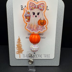 May include: A Halloween-themed badge reel featuring a cute ghost character holding a pumpkin and a beverage. The badge reel has an orange pumpkin accent and a decorative beaded element. The text "HANDMADE WITH LOVE" is at the top.