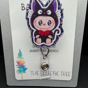 May include: A badge reel featuring a cartoon character in a cat costume with a red bow tie. The badge reel has a glittery purple border and the text "HANDMADE WITH LOVE" at the top. The text "THE ECLECTIC TREE" is at the bottom.