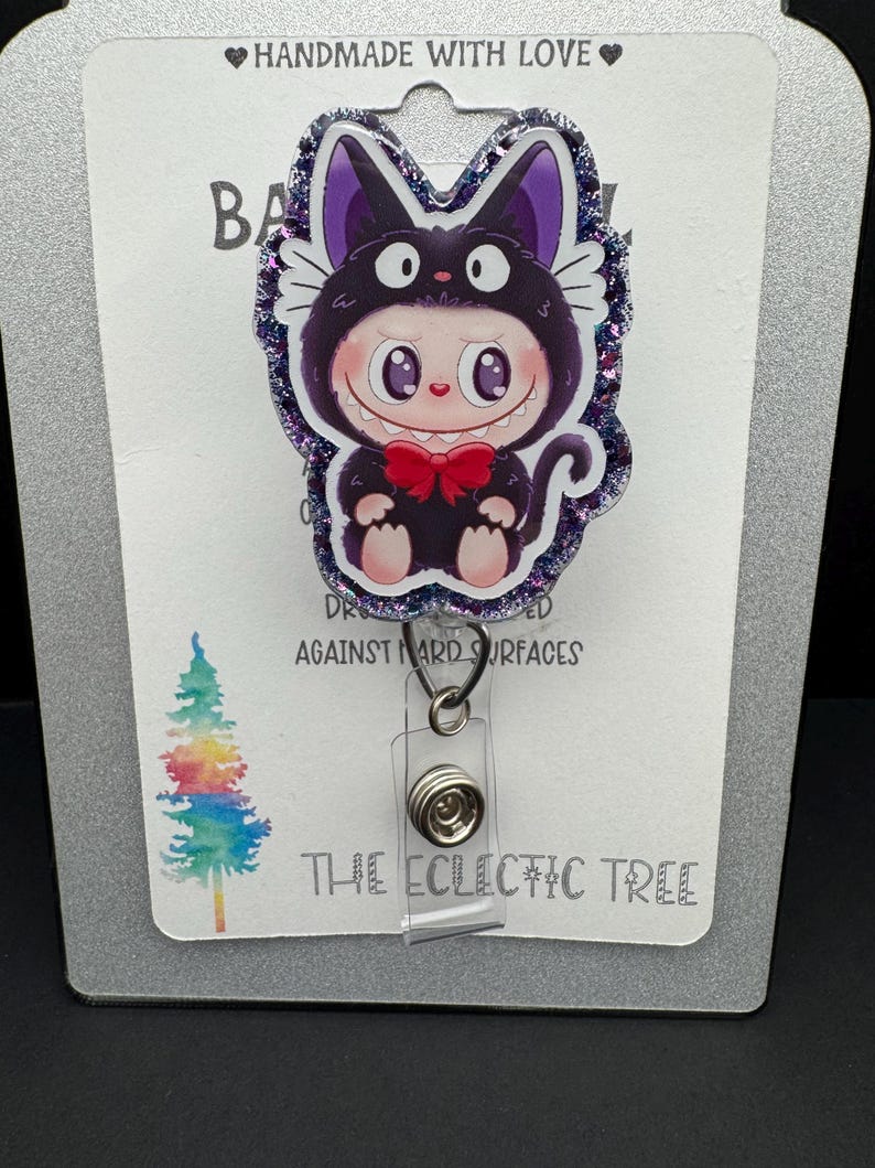 May include: A badge reel featuring a cartoon character in a cat costume with a red bow tie. The badge reel has a glittery purple border and the text "HANDMADE WITH LOVE" at the top. The text "THE ECLECTIC TREE" is at the bottom.