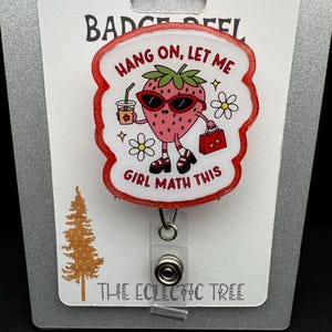 Peut inclure: Un porte-badge avec une fraise de dessin animé portant des lunettes de soleil, tenant une boisson et un sac à main. Le texte "HANG ON, LET ME GIRL MATH THIS" est affiché. Le porte-badge est attaché à une carte blanche avec le texte "HANDMADE WITH LOVE" et "THE ECLECTIC TREE".
