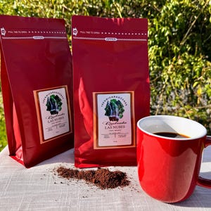 Puede incluir: Dos bolsas de café rojas y una taza roja llena de café se muestran sobre una superficie blanca. Las bolsas tienen etiquetas con texto y un gráfico de cascada. Un pequeño montón de posos de café se encuentra cerca de la taza.