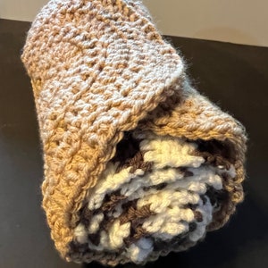 May include: A crocheted blanket in brown, white, and beige colors. The blanket is rolled up and the outer layer is a beige crocheted fabric with a scalloped edge.