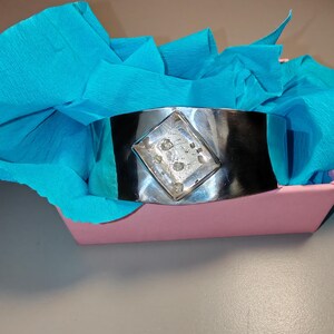 May include: A wide, silver-toned cuff bracelet with a central diamond-shaped embellishment. The bracelet is set against a backdrop of bright blue tissue paper inside a pink container. The cuff is a bold statement piece.