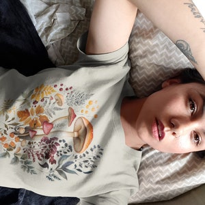 May include: A light grey t-shirt featuring a colorful illustration of mushrooms, leaves, and berries. The design includes red, orange, and brown mushrooms with detailed botanical elements. The shirt is made of a soft material.