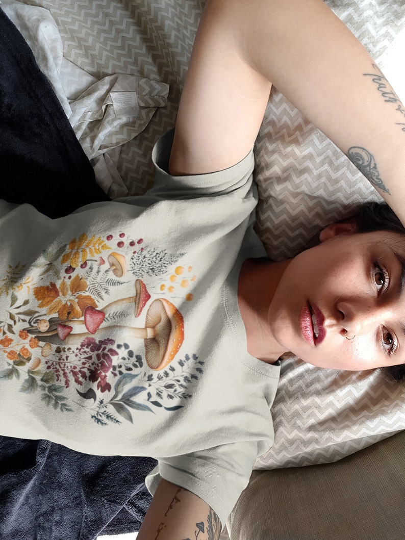 May include: A light grey t-shirt featuring a colorful illustration of mushrooms, leaves, and berries. The design includes red, orange, and brown mushrooms with detailed botanical elements. The shirt is made of a soft material.