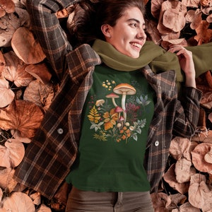 May include: Dark green t-shirt featuring a watercolor illustration of mushrooms and foliage. The shirt is worn with a brown plaid jacket, olive green scarf, and brown corduroy pants. The person is lying on a bed of autumn leaves.