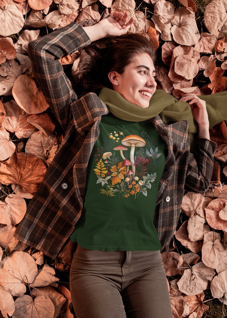 May include: Dark green t-shirt featuring a watercolor illustration of mushrooms and foliage. The shirt is worn with a brown plaid jacket, olive green scarf, and brown corduroy pants. The person is lying on a bed of autumn leaves.