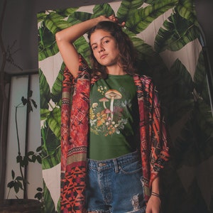 May include: A dark green t-shirt featuring a watercolor illustration of mushrooms and foliage. The model is wearing denim shorts and a patterned kimono. The background includes a tropical leaf print.