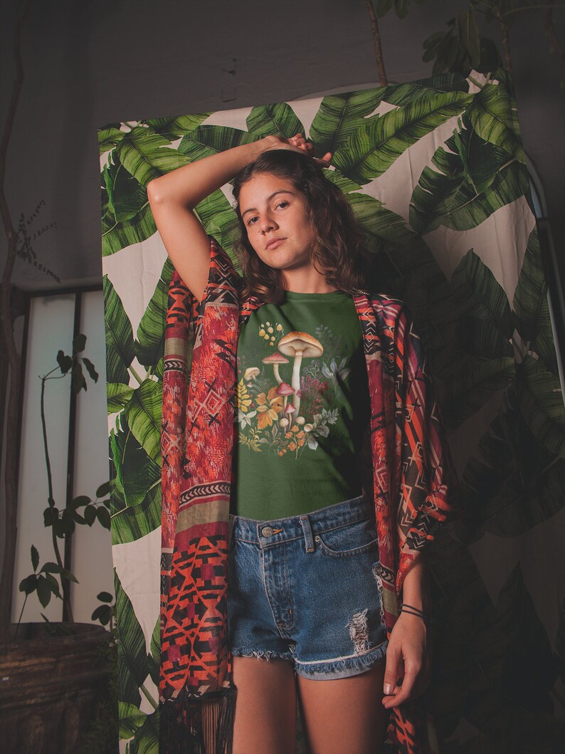 May include: A dark green t-shirt featuring a watercolor illustration of mushrooms and foliage. The model is wearing denim shorts and a patterned kimono. The background includes a tropical leaf print.