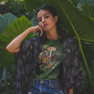 May include: Green t-shirt featuring a mushroom design with various fungi and foliage. The model is wearing denim shorts and a patterned kimono. The background includes large green leaves and a blurred natural setting.