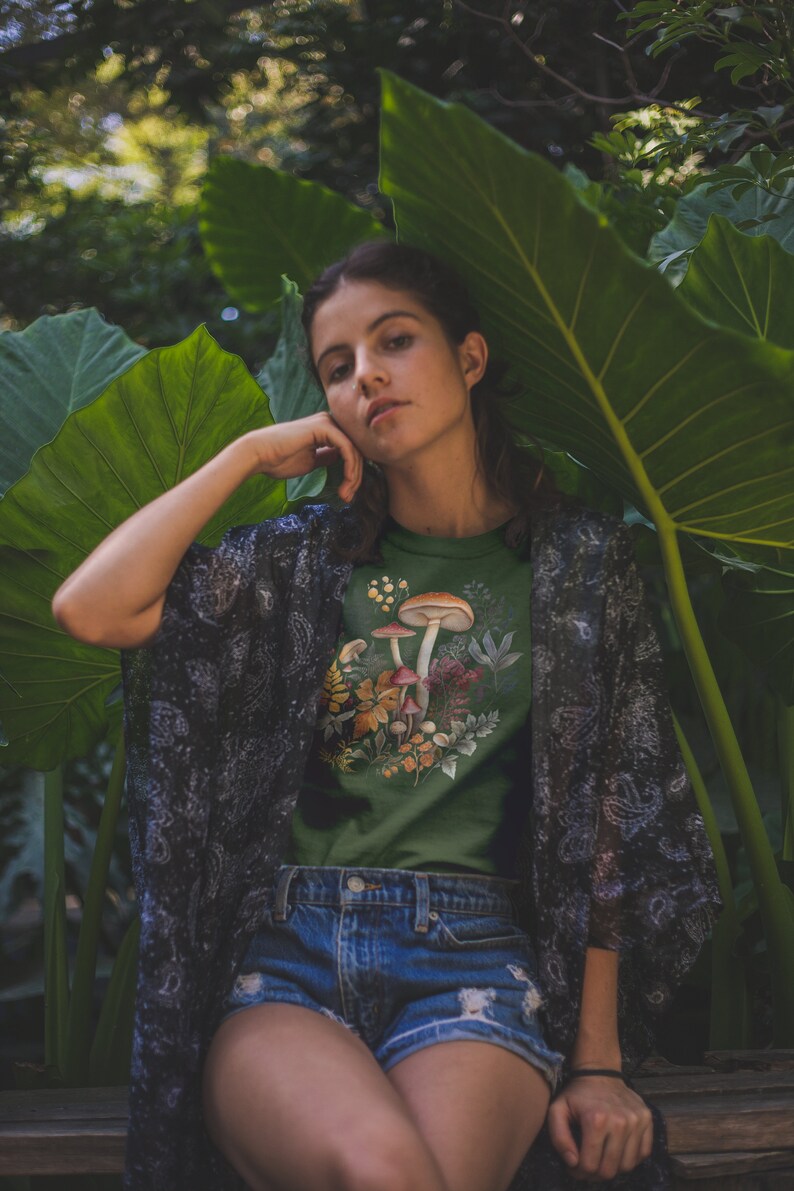 May include: Green t-shirt featuring a mushroom design with various fungi and foliage. The model is wearing denim shorts and a patterned kimono. The background includes large green leaves and a blurred natural setting.