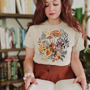 May include: Beige t-shirt featuring a colorful mushroom and floral design. The graphic includes orange, brown, and red mushrooms surrounded by various plants and leaves. The shirt is short-sleeved and made of a soft material.