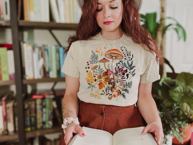 May include: Beige t-shirt featuring a colorful mushroom and floral design. The graphic includes orange, brown, and red mushrooms surrounded by various plants and leaves. The shirt is short-sleeved and made of a soft material.
