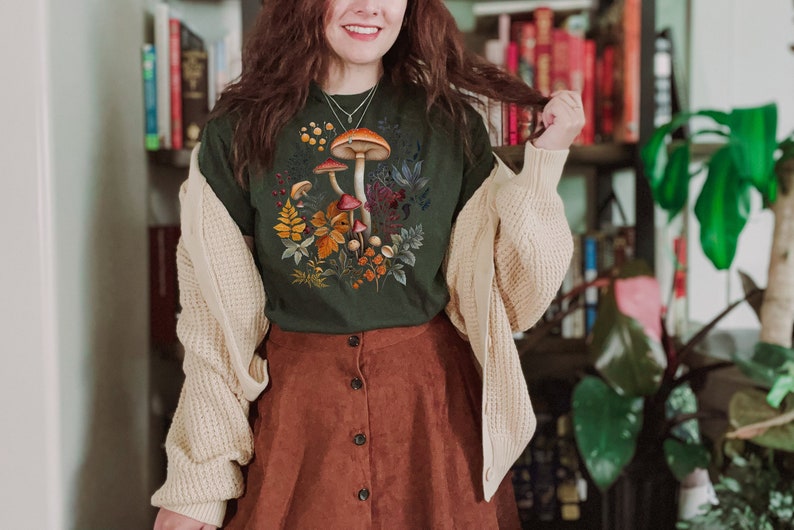 May include: Dark green t-shirt featuring a colorful mushroom and foliage design. The shirt is paired with a brown button-down skirt and a cream-colored cardigan. The image showcases a fall-themed aesthetic, with a focus on nature-inspired apparel.