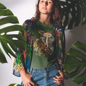 May include: An olive green t-shirt featuring a mushroom graphic, paired with blue jeans and a floral patterned kimono. The image is framed by large green leaves, creating a natural, botanical theme.