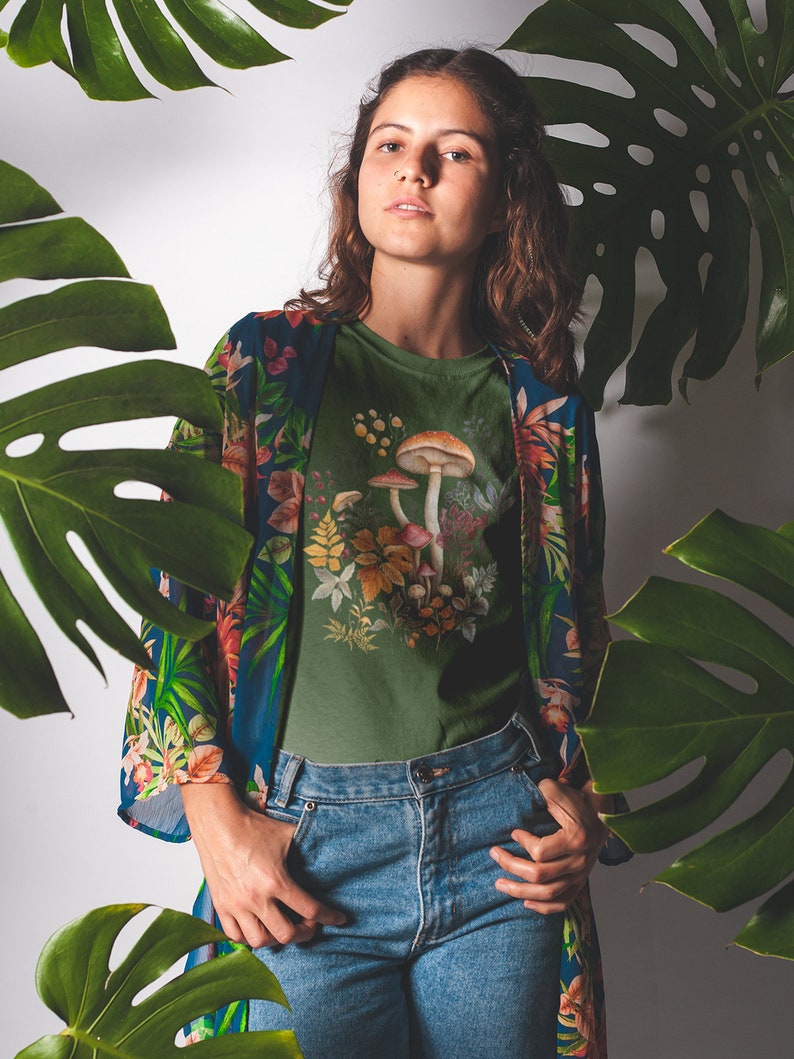 May include: An olive green t-shirt featuring a mushroom graphic, paired with blue jeans and a floral patterned kimono. The image is framed by large green leaves, creating a natural, botanical theme.