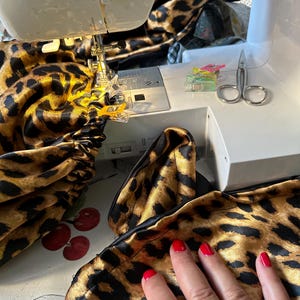 May include: A close-up of a sewing machine with a leopard print fabric being sewn. The fabric is being held in place with clips and the needle is visible. A hand is resting on the fabric.