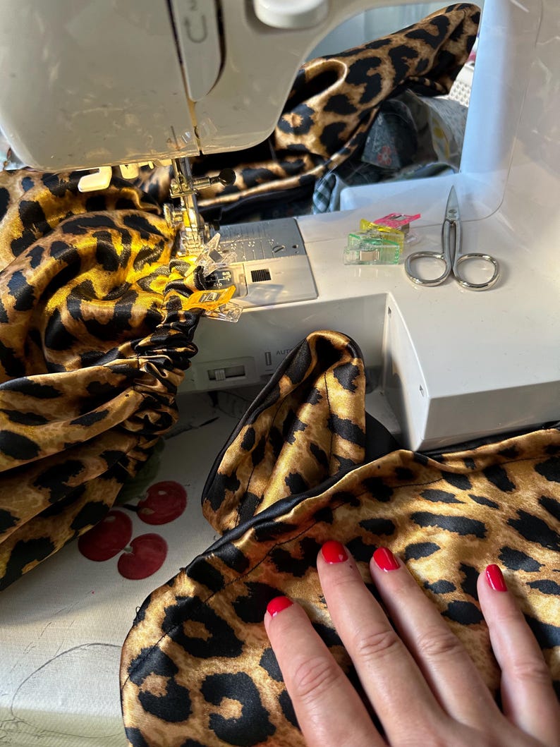 May include: A close-up of a sewing machine with a leopard print fabric being sewn. The fabric is being held in place with clips and the needle is visible. A hand is resting on the fabric.