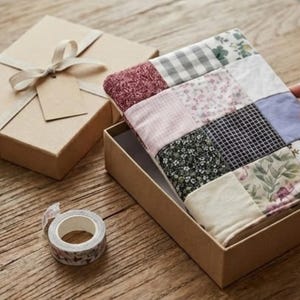 Pode incluir: Um artigo de tecido patchwork retangular com vários padrões, incluindo flores, xadrez vichy e cores sólidas em tons de rosa, verde, preto e branco, colocado dentro de uma caixa de papelão marrom ao lado de uma caixa de presente com fita e fita adesiva.