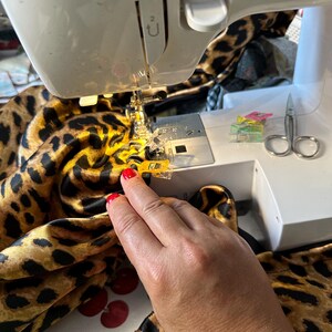 May include: A close-up of a sewing machine with a leopard print fabric being sewn. The fabric is being held in place with yellow clips. A pair of scissors and more clips are visible on the table.