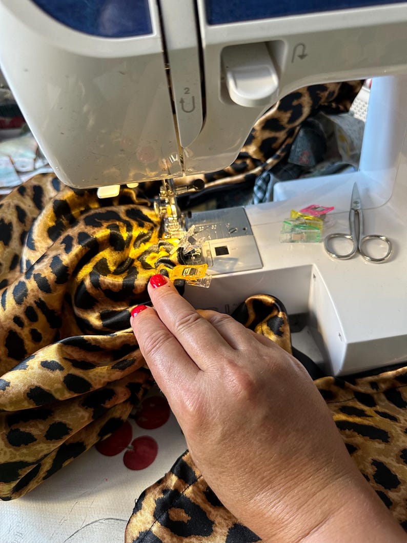 May include: A close-up of a sewing machine with a leopard print fabric being sewn. The fabric is being held in place with yellow clips. A pair of scissors and more clips are visible on the table.