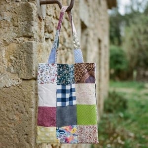 Pode incluir: Uma sacola tote de patchwork com uma variedade de padrões de tecido, incluindo floral, bolinhas e xadrez. A sacola está pendurada em um gancho de metal em uma parede de pedra. A alça da sacola é feita de tiras de tecido coordenadas.