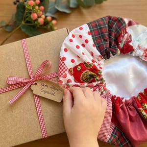 Pode incluir: Uma caixa de presente de papelão marrom amarrada com uma fita xadrez vermelha e branca e uma etiqueta com a inscrição Para Alguém Especial, ao lado de uma touca de cabelo circular com patchwork de padrões vermelhos, brancos e verdes e forro de cetim.